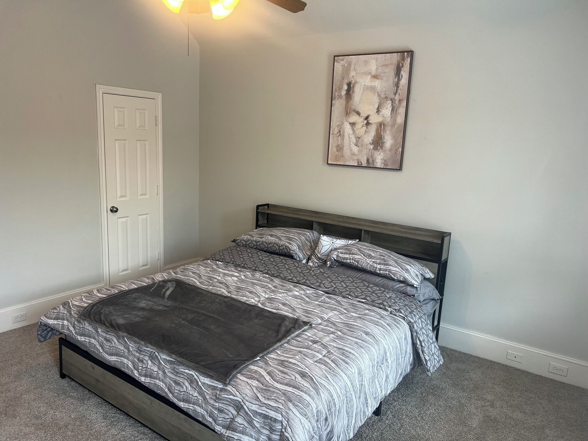 A bedroom with a bed, a door, a ceiling fan, and a wall-mounted abstract painting.