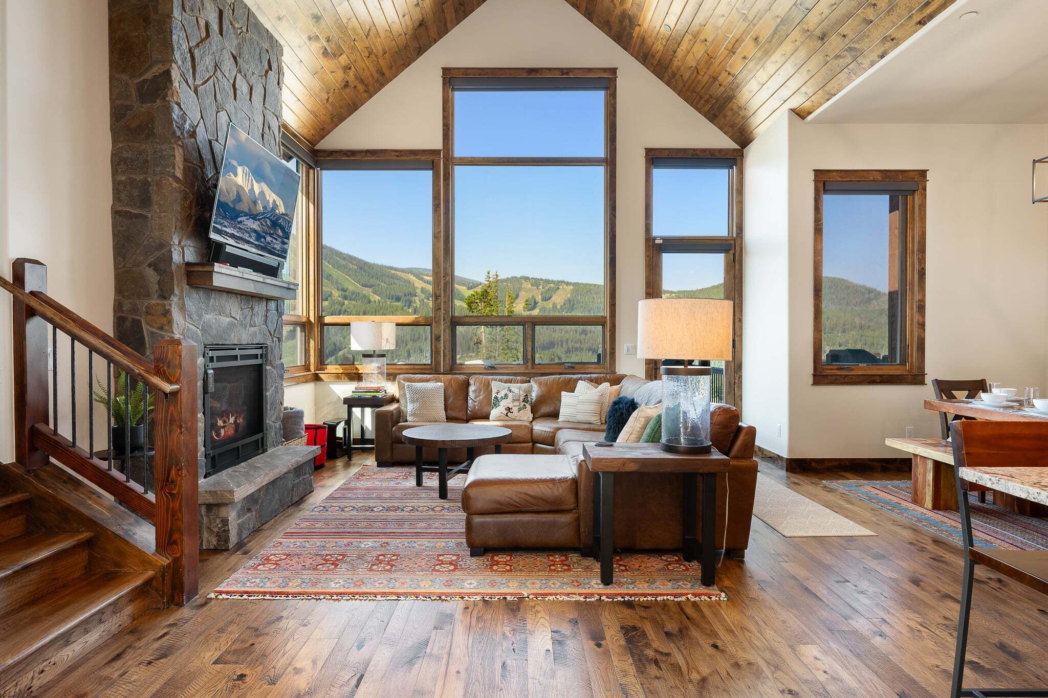 A spacious living room with a stone fireplace, large windows, and a wooden staircase.
