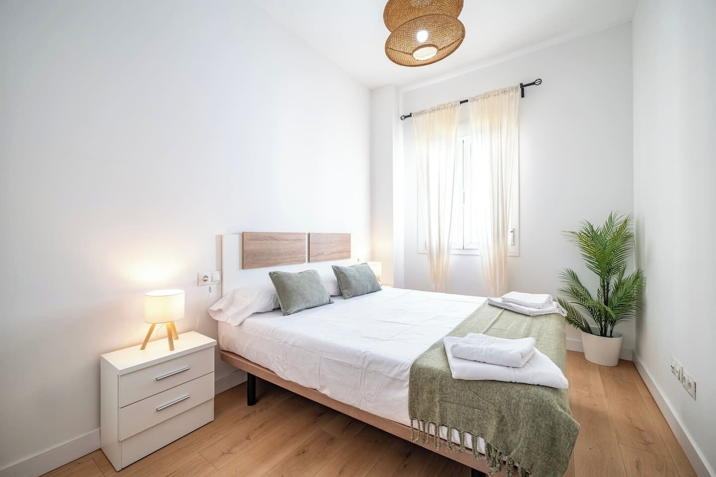 Een netjes opgemaakt bed met een houten hoofdeinde, een bedlampje, een klein nachtkastje met lades, een groene plaid en een pot met een plant.