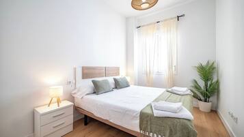 Een netjes opgemaakt bed met een houten hoofdeinde, een bedlampje, een klein nachtkastje met lades, een groene plaid en een pot met een plant.