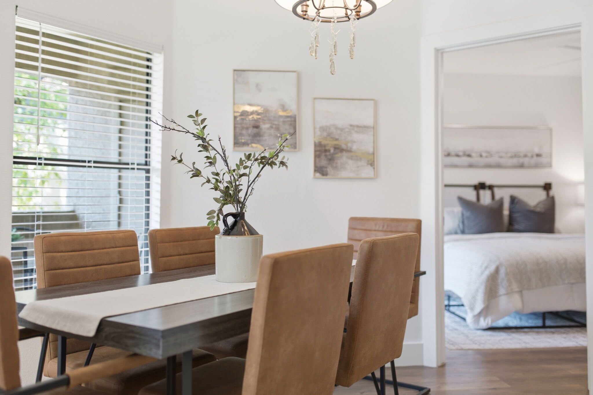 Uma área de refeições com uma mesa, cadeiras e um vaso com galhos. Há quadros nas paredes e uma cama ao fundo.