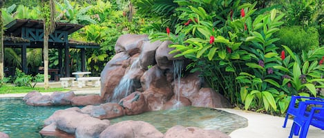 A rock waterfall in a tropical garden with a pool, benches, and a gazebo.