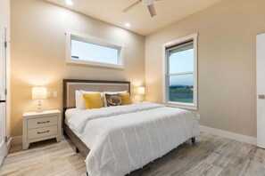 Un dormitorio con cama, mesitas de noche, ventilador de techo y dos ventanas.