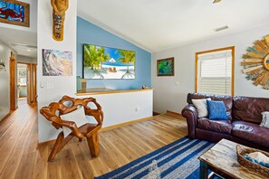 A living room with a wooden floor, a brown leather sofa, a wooden chair, a blue-striped rug, and a television on a blue wall. - Till Next Time - hot tub, tiki bar, EV charger, and beach access. (Kill Devil Hills)
