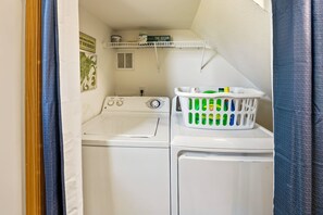 A laundry room with a front-loading washer and dryer, a shelf with cleaning supplies, and a laundry basket with detergent bottles. - Till Next Time - hot tub, tiki bar, EV charger, and beach access. (Kill Devil Hills)