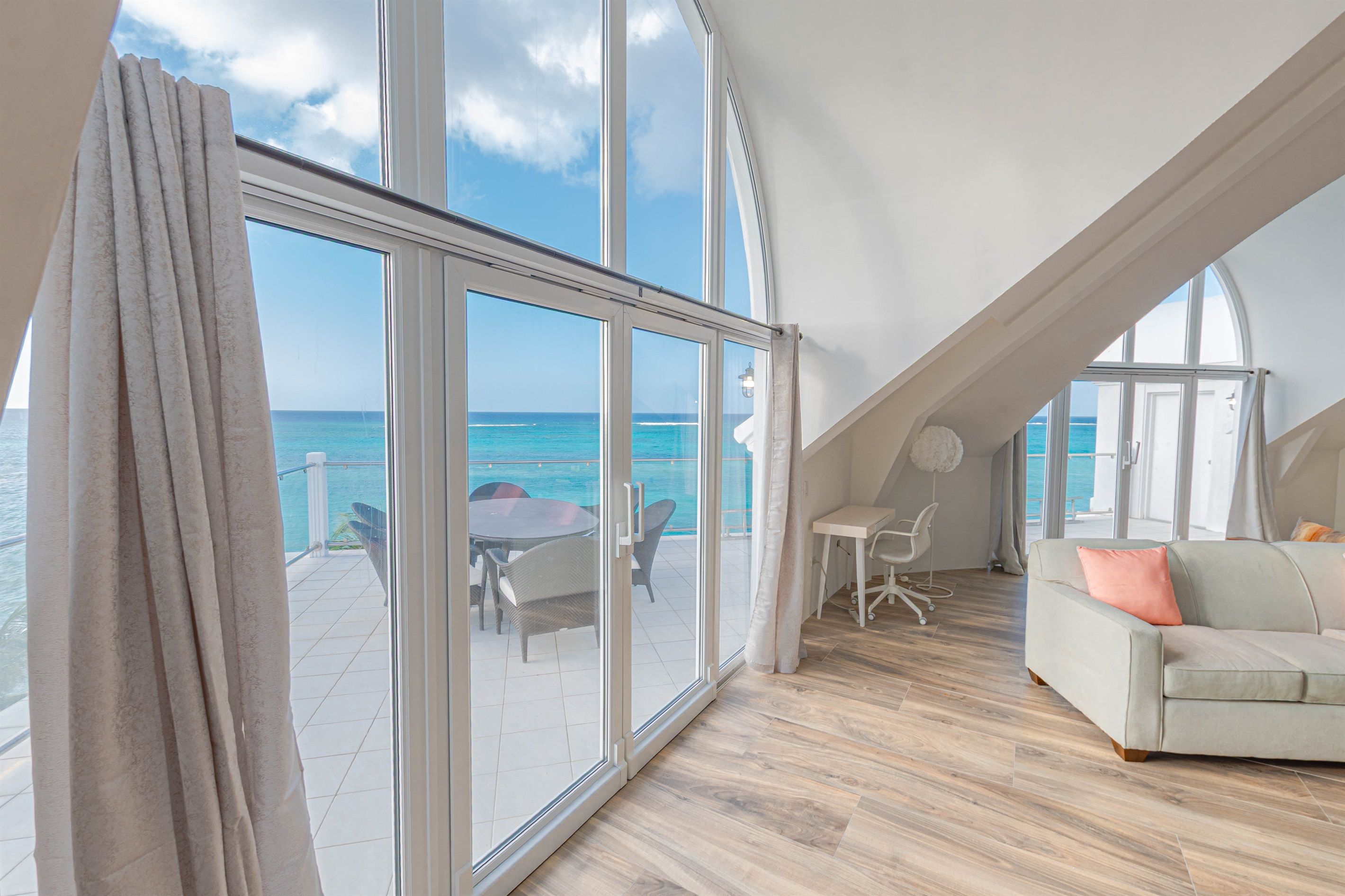Amplia habitación con un gran ventanal corredero que da a un balcón con vistas al mar. Hay un sofá blanco y una pequeña mesa con una silla en la habitación.