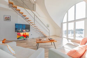 A modern living room with a wooden staircase, a flat-screen TV, a colorful cabinet, and a large window offering a view. - Love Beach W4 - Penthouse Steps to Pool & Beach (Nassau)
