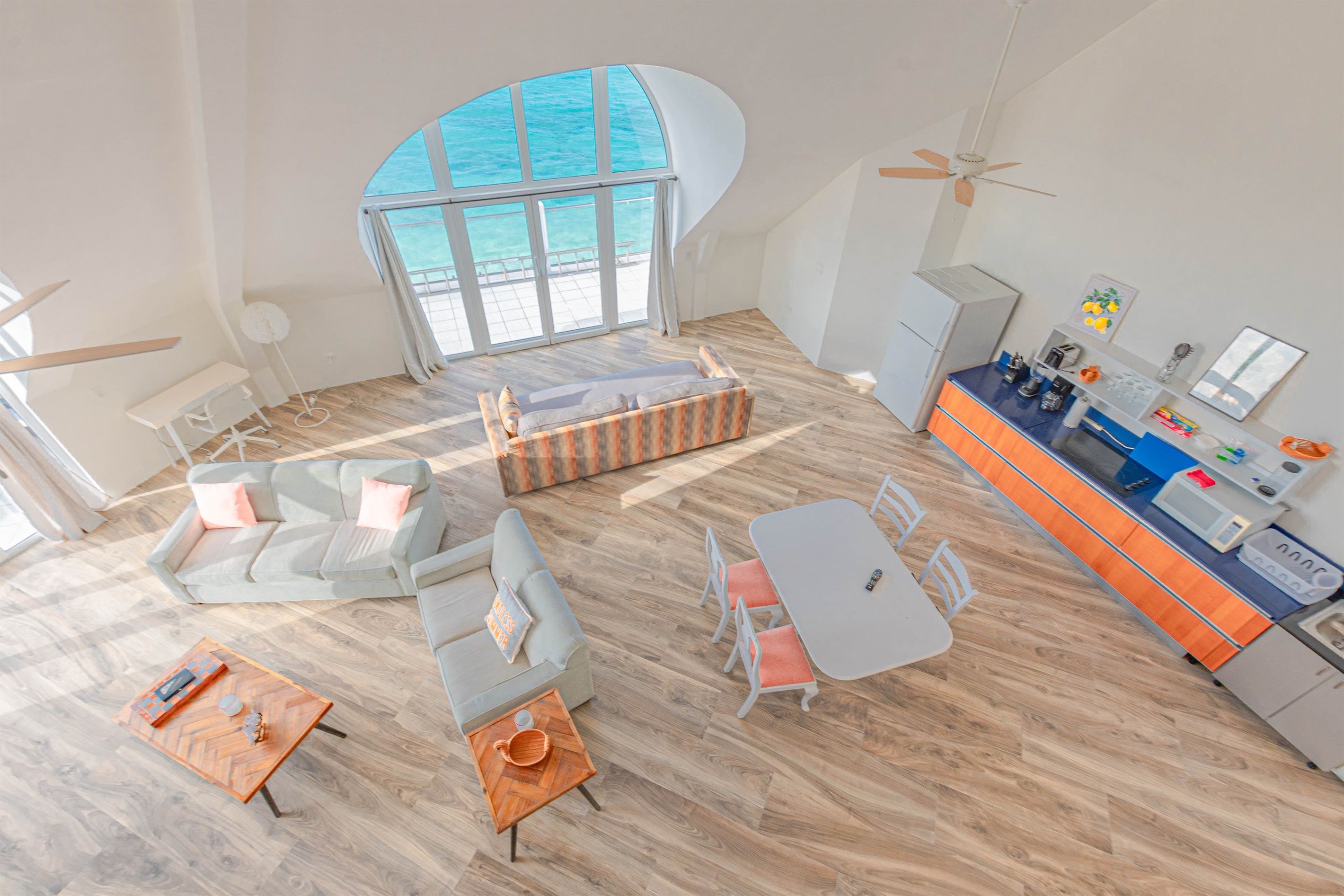 A modern interior with a large curved window overlooking the sea, a kitchen area with a blue countertop, and a seating area with a sofa and a coffee table.
