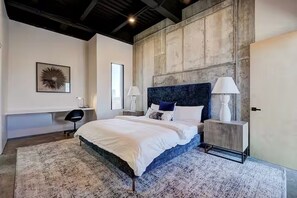 A modern bedroom with a large bed, bedside lamps, a bench, and a wall-mounted artwork. - TB Signature Lofts – Unit 210 in Downtown OKC (Oklahoma City)