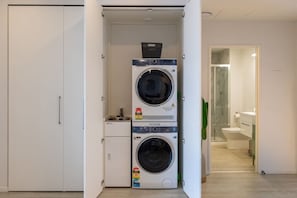 A compact laundry room with two stacked washing machines, a small sink, and a toilet visible through an open door. - Heart of CBD - Walk to ONE NZ Stadium + Parking (Christchurch)