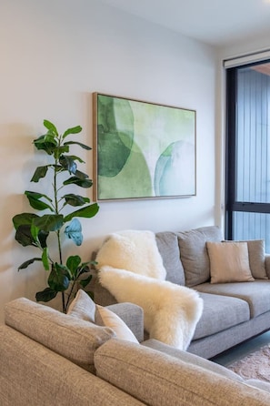 A living room with a grey sofa, a white fluffy throw, and a green abstract painting on the wall. - Heart of CBD - Walk to ONE NZ Stadium + Parking (Christchurch)