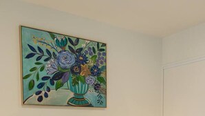 Ett hotellrum med en säng, lila kuddar, ett grönt och vitt randigt sängöverkast, en lila pläd och en tavla med blommor på väggen.