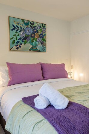 A hotel room with a bed, purple pillows, a green and white striped bedspread, a purple throw blanket, and a painting of flowers on the wall. - Ultimate City Comfort - Carpark + EV (Christchurch)