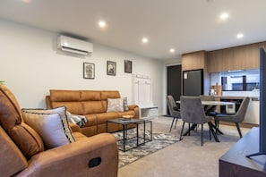 A modern living room with a sofa, a coffee table, and a dining area with chairs and a table. - Ultimate City Comfort - Carpark + EV (Christchurch)