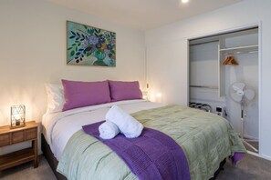 Ein Schlafzimmer mit Bett, Kissen, Decke, Nachttisch mit Lampe, Wandkunst, Kleiderschrank und Standventilator.