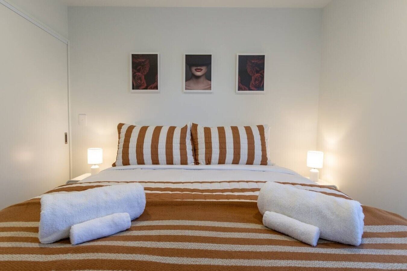 A hotel room with a bed, striped bedding, two pillows, two bedside lamps, and three framed pictures on the wall.