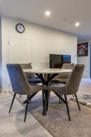 A dining area with a round table, four chairs, a wall clock, and a television. - Ultimate City Comfort - Carpark + EV (Christchurch)