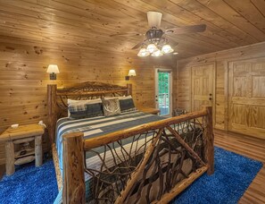 A wooden cabin bedroom with a bed, two bedside lamps, a ceiling fan, and a door with a window. - Copper Top - Mountain Views | Hot Tub | Expansive Patio (Blue Ridge)