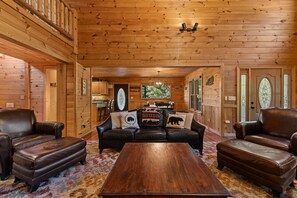A log cabin interior with a central seating area featuring a sofa and two armchairs, a wooden coffee table, and a dining table with chairs. - Copper Top - Mountain Views | Hot Tub | Expansive Patio (Blue Ridge)