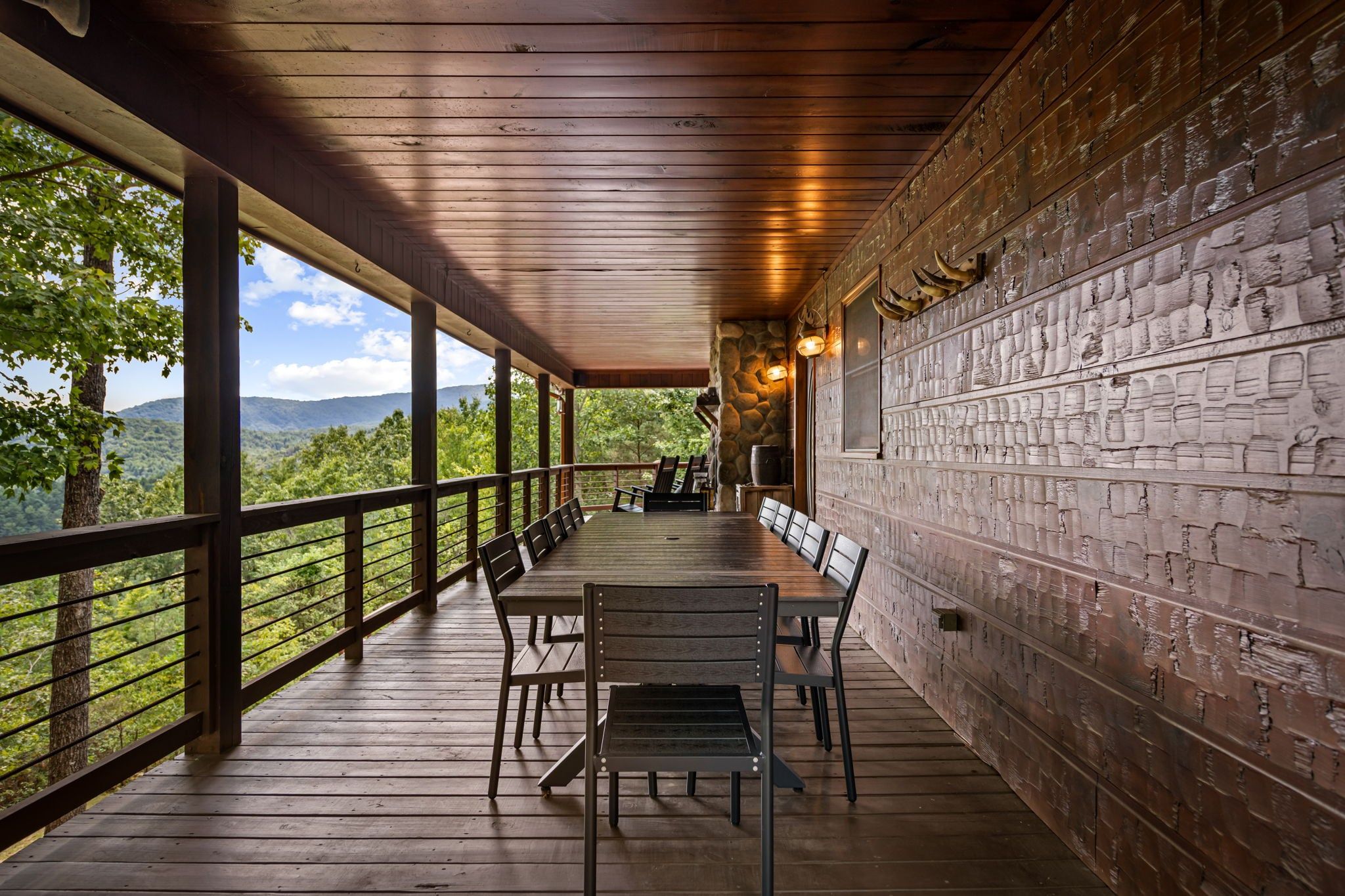 Un espacio de estar al aire libre con techo, con una mesa larga y sillas, un muro de piedra con diseños tallados y vistas a un paisaje boscoso.