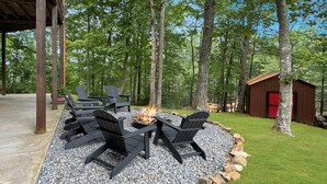 Et rundt udendørs opholdsområde med Adirondack-stole omkring et bålsted, omgivet af en kant af grus og sten, med en trækonstruktion og et skur i baggrunden.