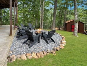 A circular outdoor seating area with Adirondack chairs around a fire pit, surrounded by a gravel and rock border, with a wooden structure and a shed in the background. - Copper Top - Mountain Views | Hot Tub | Expansive Patio (Blue Ridge)