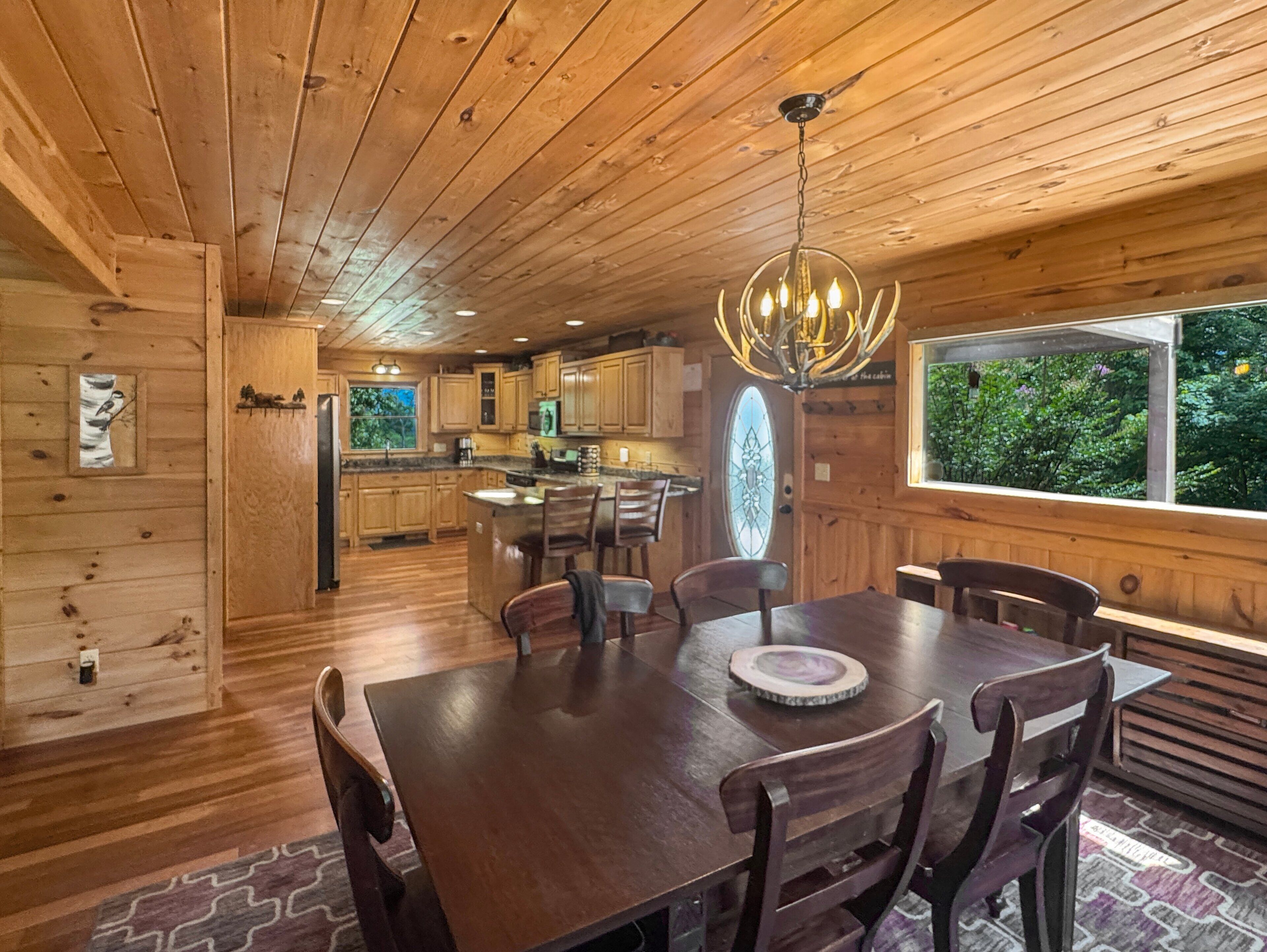 A wooden cabin interior with a dining area, kitchen, and a large window overlooking greenery.