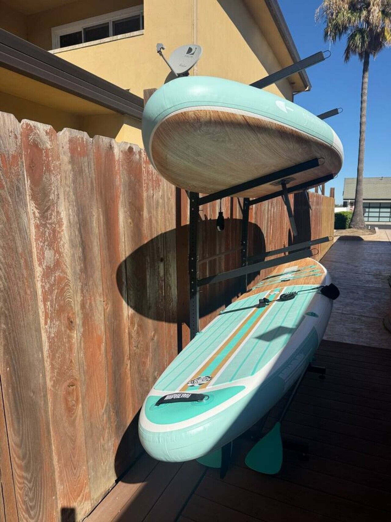 A paddleboard leaning against a wooden fence with a satellite dish on a building in the background.