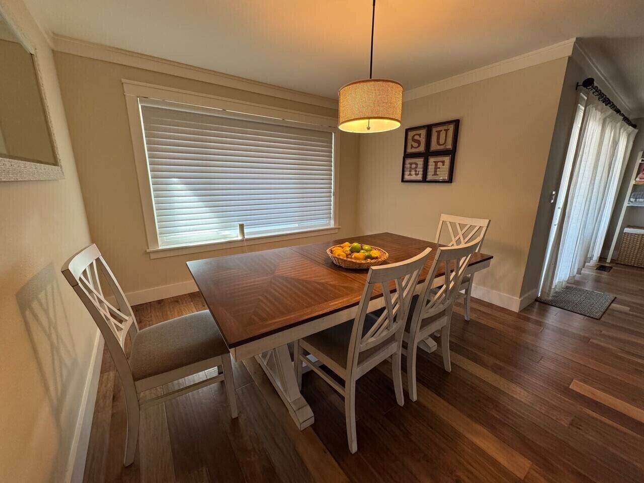 Un comedor con mesa de madera, sillas blancas, lámpara colgante y ventana con persianas.