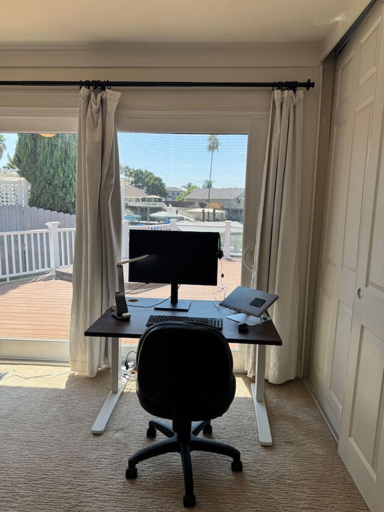 A home office with a desk, computer, and chair, overlooking a backyard with a wooden deck and a view of houses.