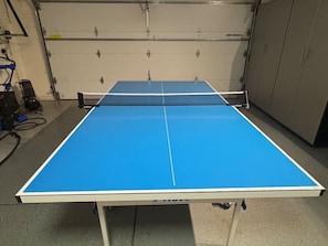 A ping pong table with a white frame and blue playing surface, located in a room with a concrete floor and metal lockers in the background.