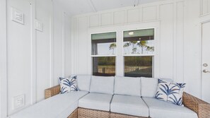 A white paneled room with a light grey sofa and two decorative pillows, a wicker chair, and a window with a view of trees and a fence. - Blue Heron Laguna Haven 253-West End-Sleeps 12 (Panama City Beach)