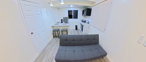 A modern interior with a grey sofa in the foreground, a kitchen area in the background, and a white door on the left.