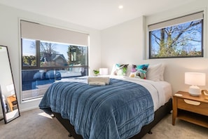 A bedroom with a large bed, a bedside table, a nightstand, and a window with a view of a residential area. - Peaceful Family CBD Stay w/ Car Park + EV charger (Christchurch)