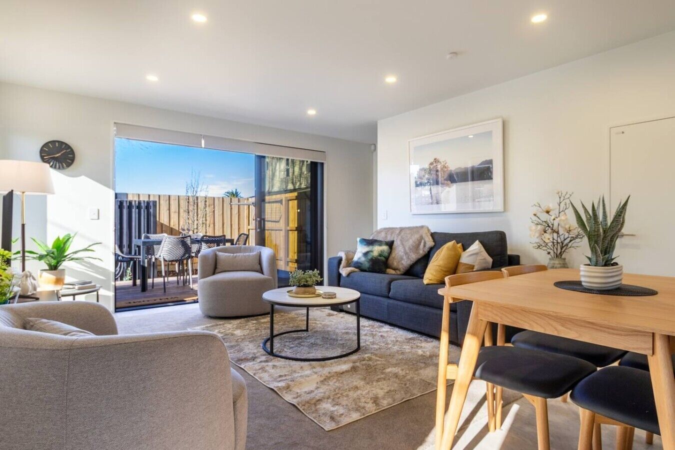 A modern living room with a sofa, coffee table, and dining area. There is a sliding glass door leading to an outdoor patio.