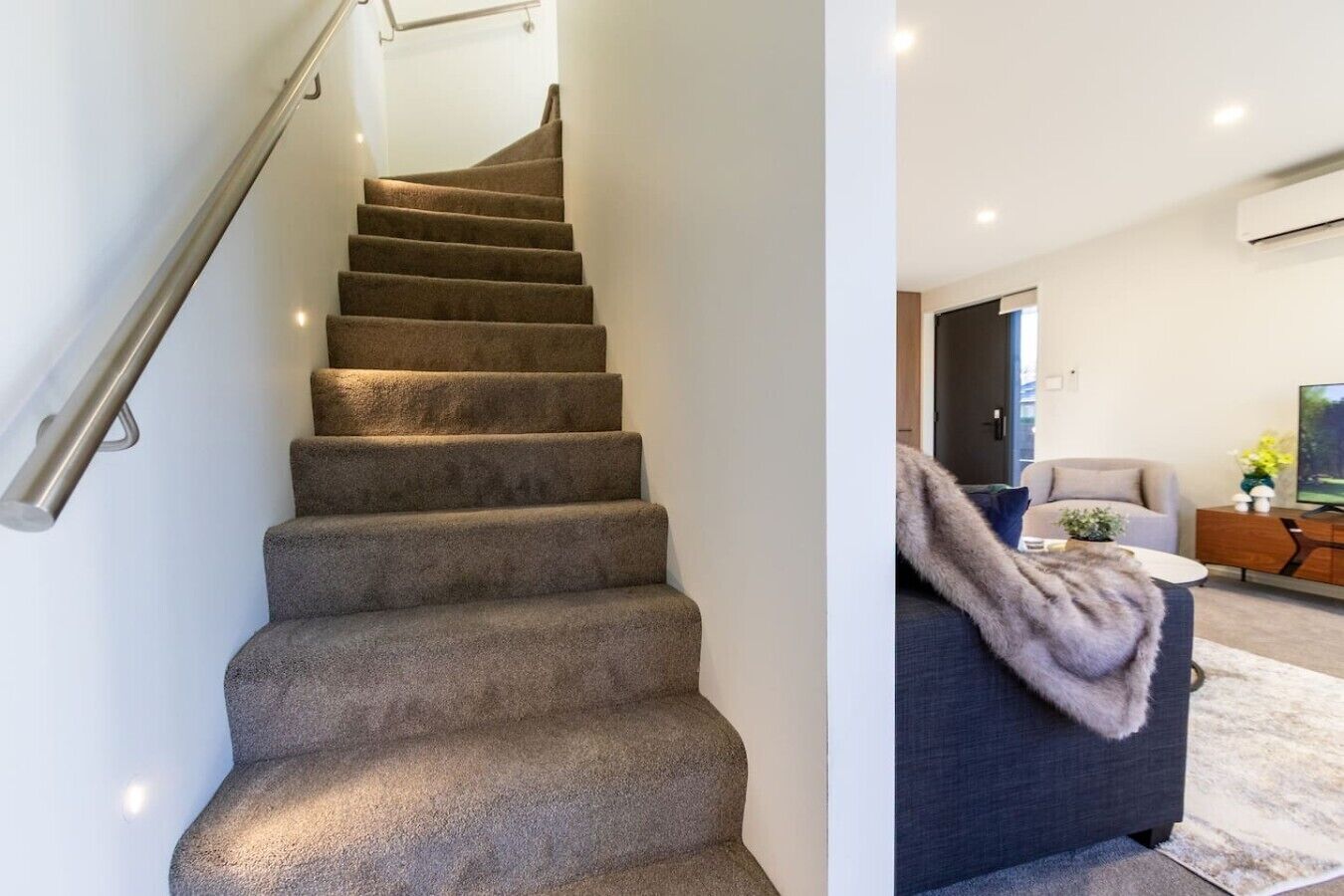 A staircase with carpeted steps and a metal handrail, leading to a living area with a sofa, a coffee table, and a television.