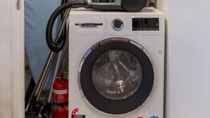 A compact laundry area with a front-loading washing machine, a vacuum cleaner, and a shelf with cleaning supplies. - Peaceful Family CBD Stay w/ Car Park + EV charger (Christchurch)
