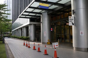 Entrada de un edificio moderno con toldo de vidrio, pilares metálicos y señal de zona de bajada.