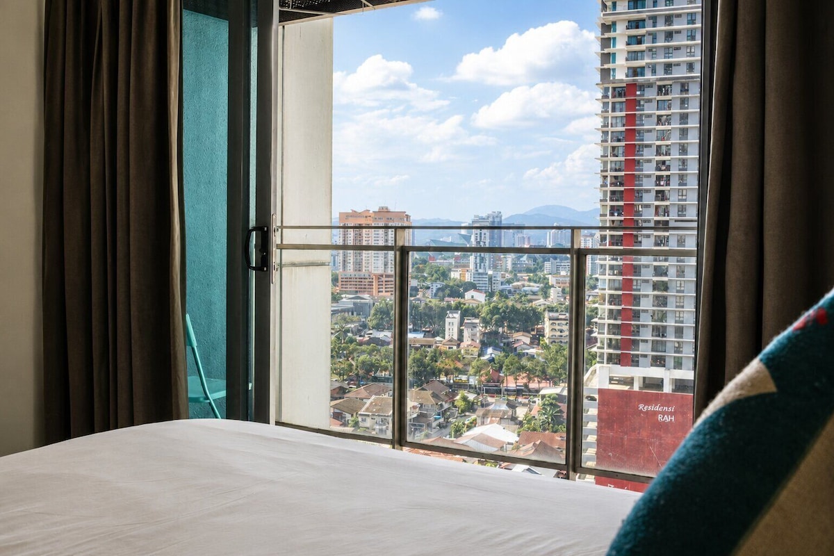 Habitación de hotel con una cama, vista de un paisaje urbano a través de un ventanal y cortinas.