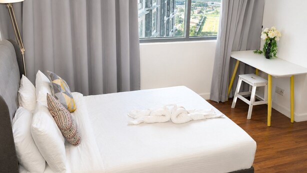 Una cama bien tendida con sábanas blancas y almohadones decorativos, un escritorio con una silla, y una ventana con vistas a edificios.