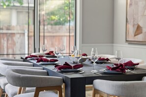 Mesa de jantar posta com pratos, talheres e copos, rodeada por cadeiras estofadas.