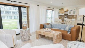 Una sala de estar moderna con sofá, mesa de centro y sillones, una cocina al fondo y ventanas amplias con cortinas.