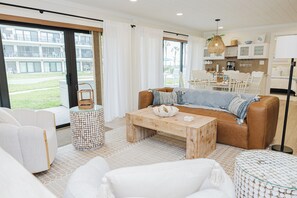 A modern living room with a sofa, coffee table, and chairs, a kitchen in the background, and large windows with curtains. - Thoughtfully Redesigned Beachside Condo (St. Augustine)