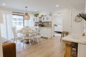 A modern kitchen with white cabinetry, a dining area with white chairs, and a view of the outside. - Thoughtfully Redesigned Beachside Condo (St. Augustine)