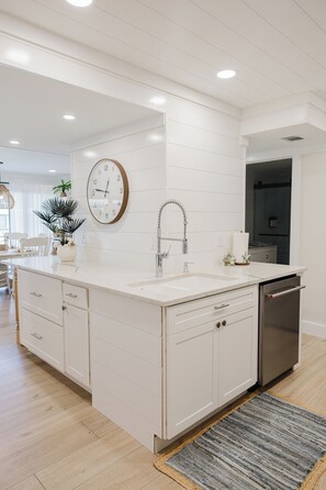 A modern kitchen with white cabinetry, a central island, a built-in oven, and a clock on the wall. - Thoughtfully Redesigned Beachside Condo (St. Augustine)