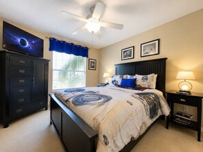 Een slaapkamer met een bed, een plafondventilator, een televisie, een raam met jaloezieën en een nachtkastje met een lamp.