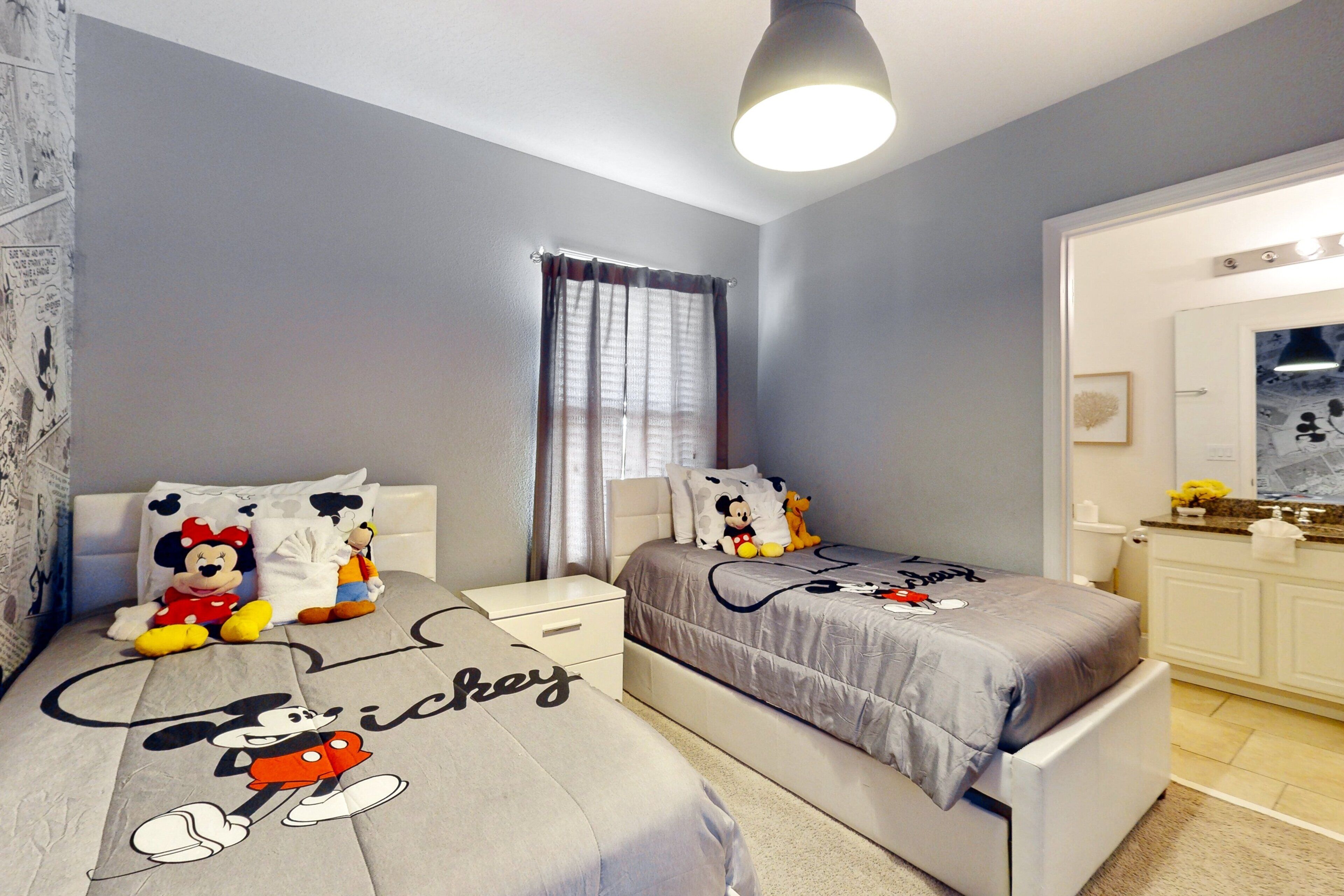 A bedroom with two beds, each with Mickey Mouse themed bedding. There is a window with curtains, a bedside table, and a nightstand.