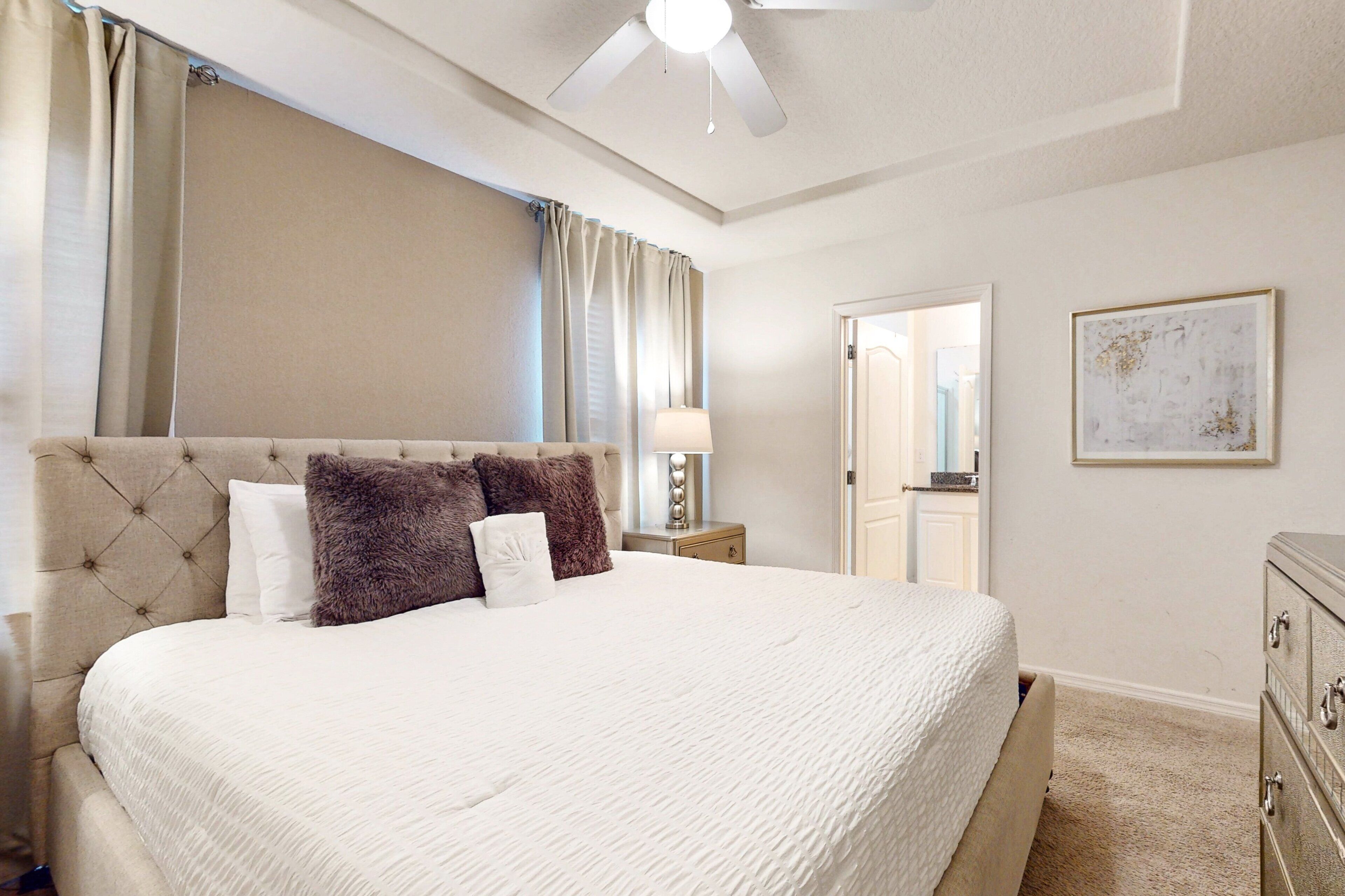 Une chambre à coucher avec un grand lit, un ventilateur de plafond, une table de chevet, une commode et une porte donnant sur une autre pièce.