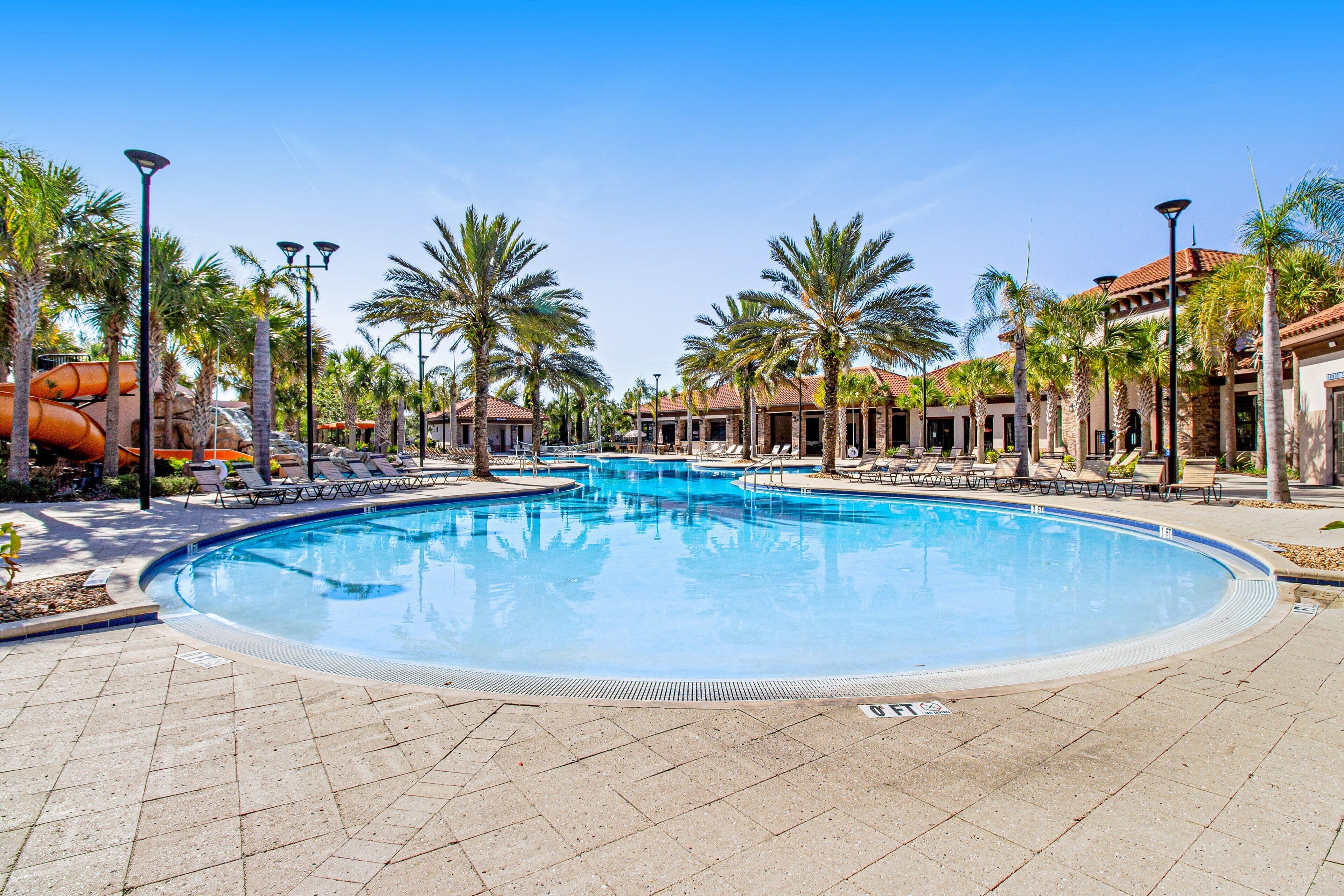 A large outdoor swimming pool surrounded by palm trees and lounge chairs, with a building in the background.