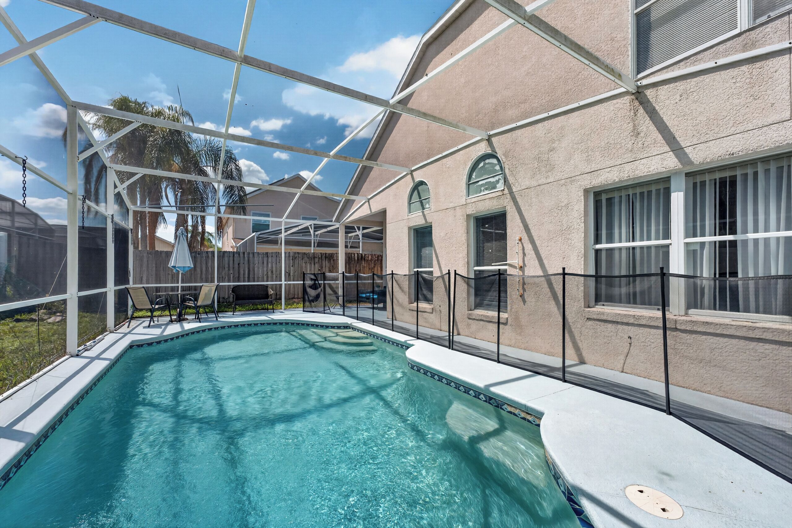 A swimming pool with a clear blue water surface, surrounded by a covered patio area with chairs and a table.